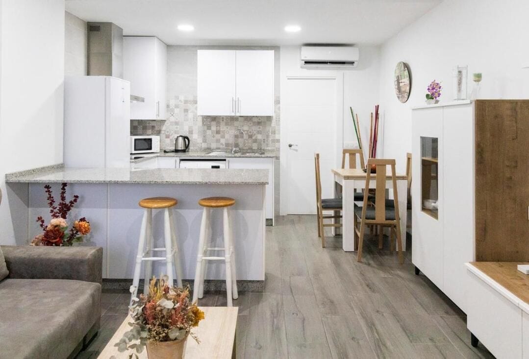 Una cocina moderna con armarios blancos, una isla central con taburetes, un comedor con sillas de madera y un sofá gris.
