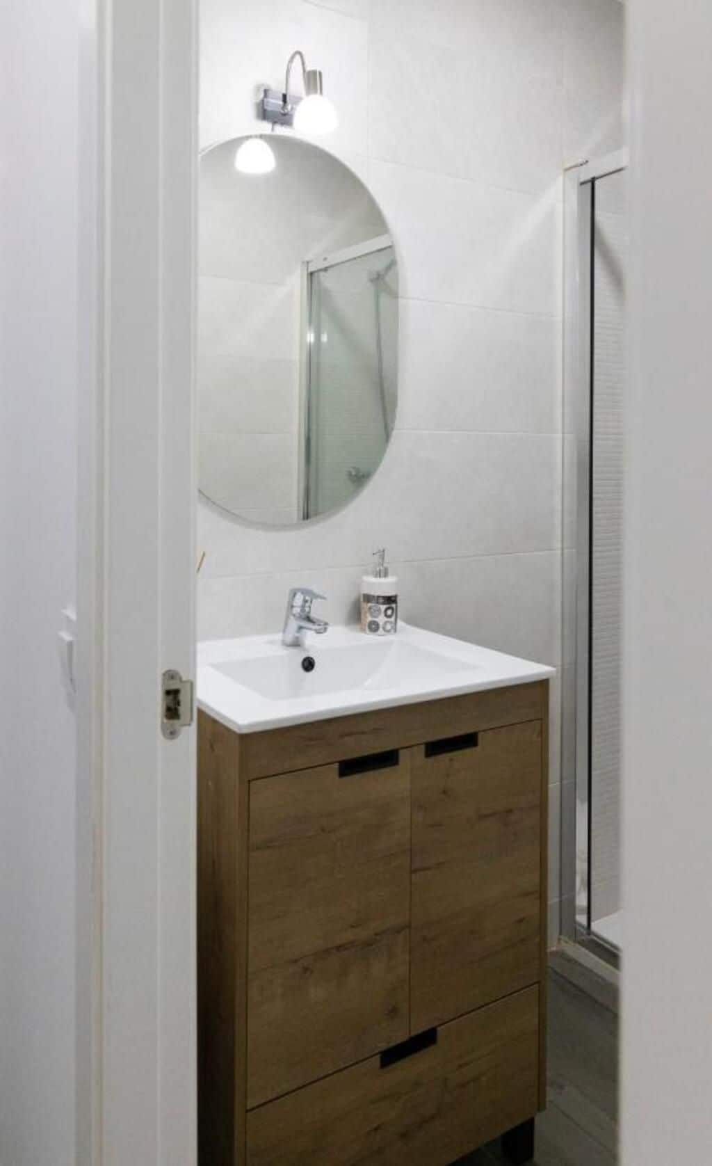 Un baño con lavamanos blanco, tocador de madera, espejo redondo y cabina de ducha con mampara de vidrio.
