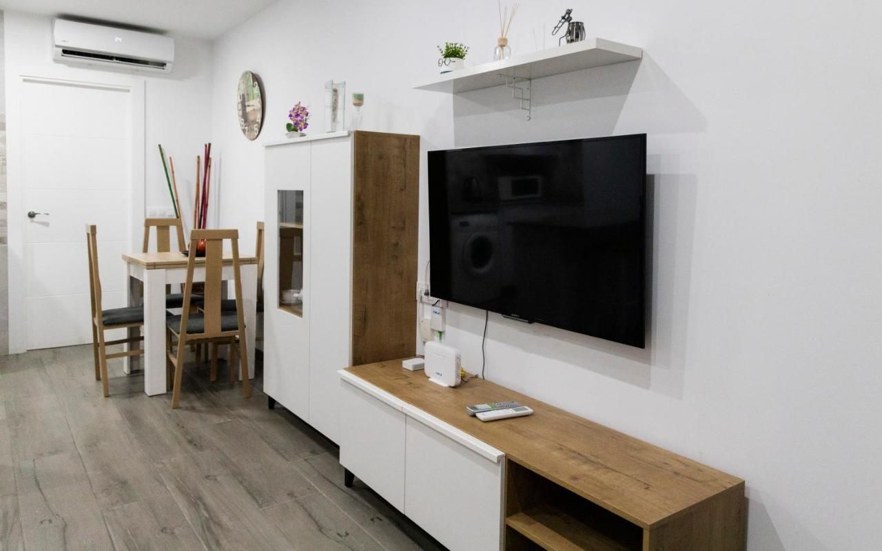 Una sala de estar con una televisión de pantalla plana montada en la pared, una mesa de centro de madera, un armario blanco y un comedor con mesa y sillas de madera.