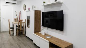 Una sala de estar con una televisión de pantalla plana montada en la pared, una mesa de centro de madera, un armario blanco y un comedor con mesa y sillas de madera.