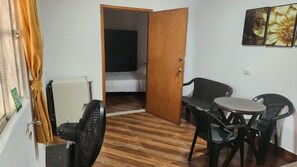 Una habitación pequeña con cama, una mesa y sillas, un ventilador y una puerta que da a otra habitación.