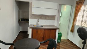 Una cocina compacta con un lavamanos, armarios de madera, una pequeña mesa de comedor y un ventilador.