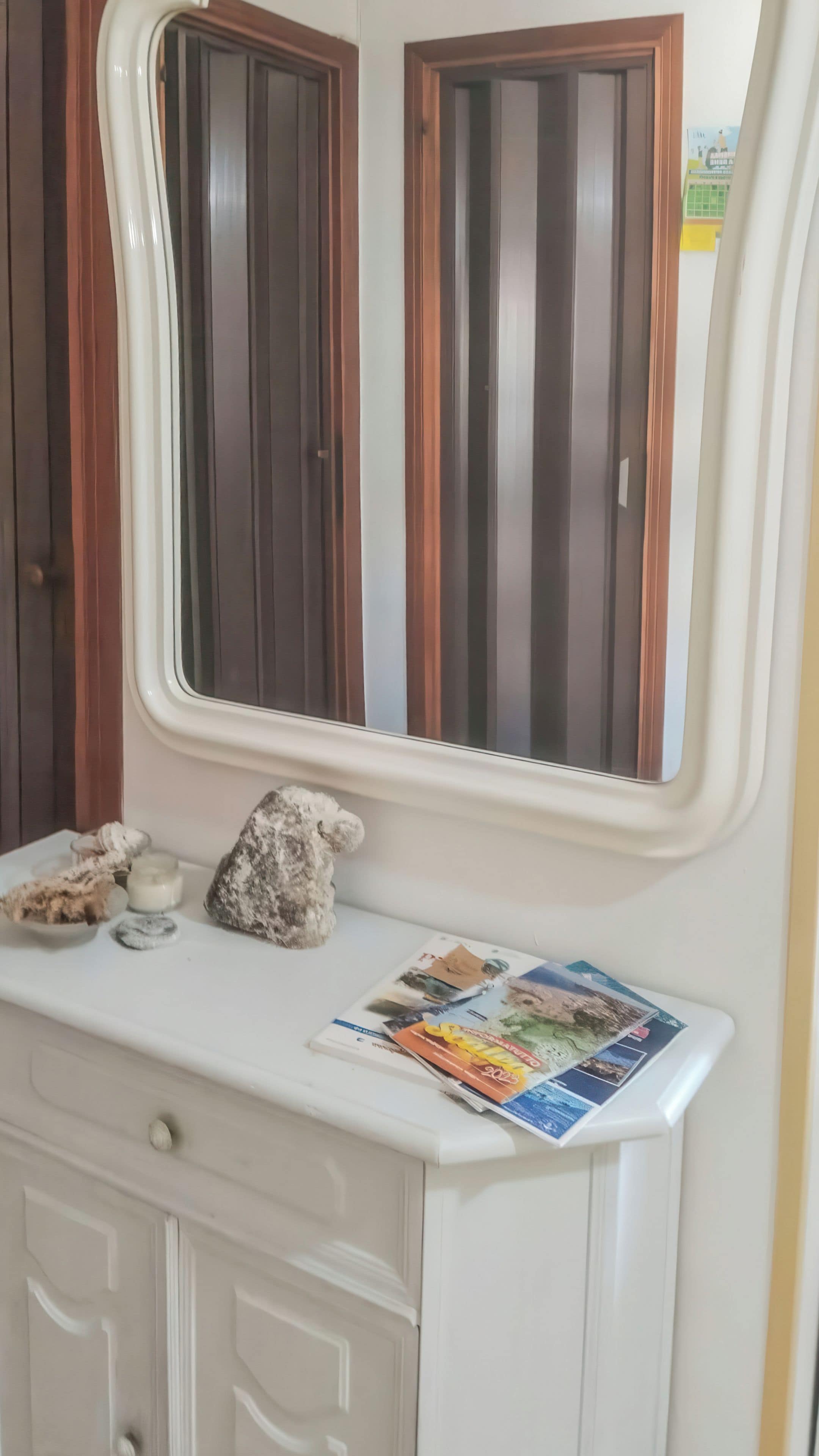 Een witte kaptafel met een spiegel erboven, een klein tafeltje met een teddybeer en een kaars, en een tijdschrift op de kaptafel.