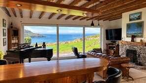 A living room with a piano, a bar area, a fireplace, and a large window overlooking a scenic landscape. - Atlantic Golf Retreat near Waterville (Cahersiveen)