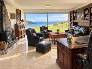 A spacious living room with a stone fireplace, leather furniture, a large window overlooking the sea, and a bookshelf filled with books and decorative items. - Atlantic Golf Retreat near Waterville (Cahersiveen)