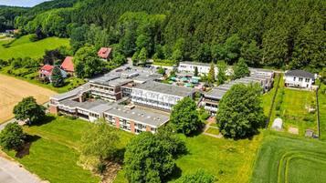 An aerial view of a campus with multiple buildings, green fields, and a forested hill in the background.