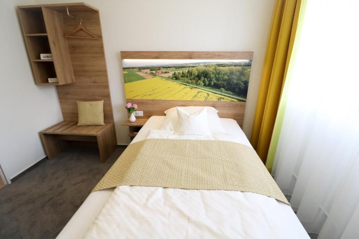 Quarto de hotel com cama, prateleira de madeira com um objeto pendurado, pequena mesa com um vaso e um grande quadro de paisagem na parede.