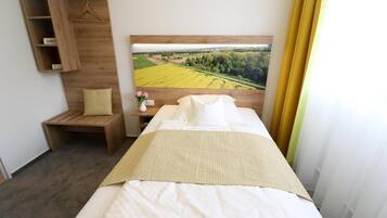 A hotel room with a bed, a wooden shelf with a hanging item, a small table with a vase, and a large landscape painting on the wall.