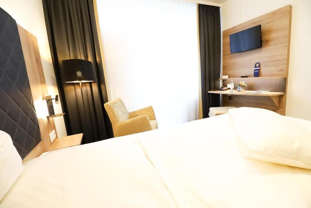 A hotel room with a bed, a desk with a flat-screen TV, a chair, and a lamp.