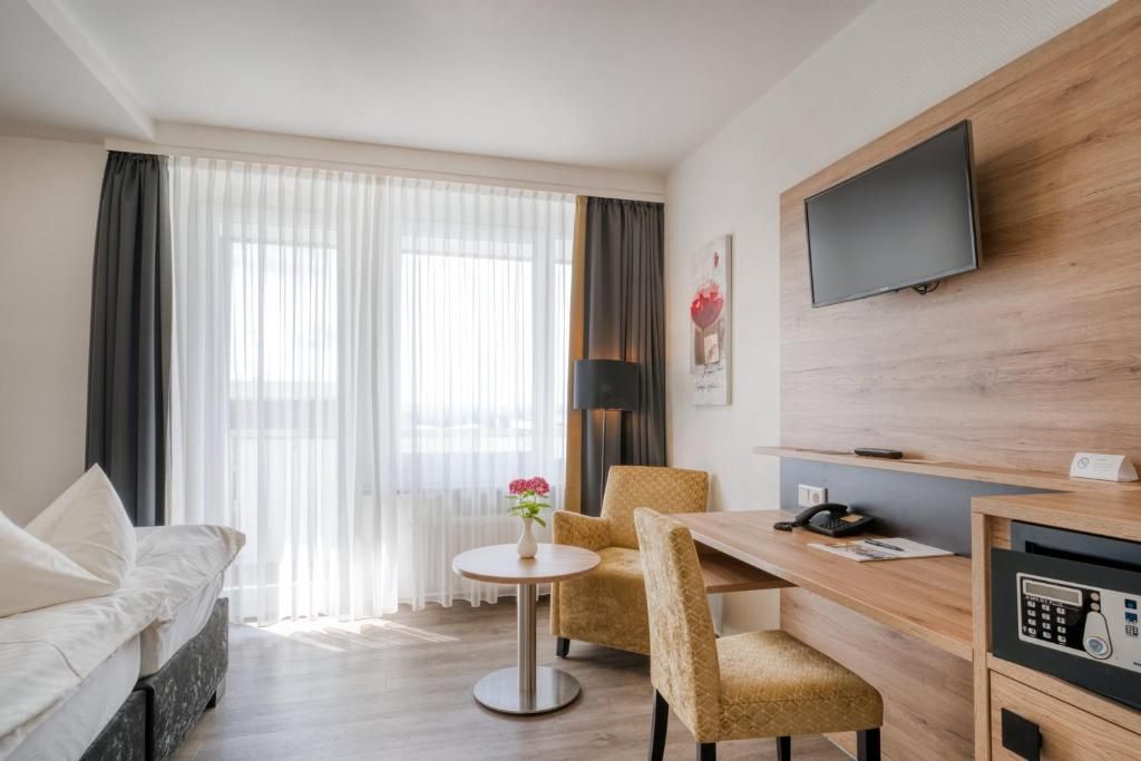 Une chambre d’hôtel comprenant un lit, une table ronde, deux chaises, un bureau avec un téléphone, un téléviseur à écran plat fixé au mur et une fenêtre avec des rideaux légers.