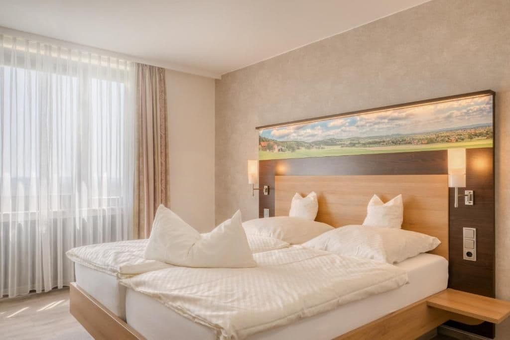 Camera d'albergo con un letto grande, una testiera con luci a parete, un quadro sopra il letto e una finestra con tende leggere.