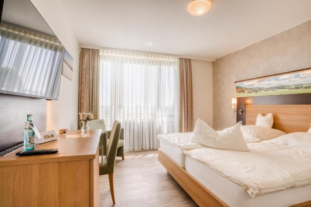 A hotel room with a bed, a desk with a chair, a TV, and a window with curtains.