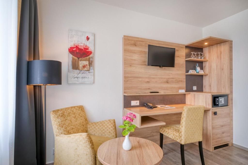 Ein modernes Zimmer mit einem an einer Holzwand befestigten Flachbildfernseher, einem kleinen runden Tisch mit Blumen in einer Vase, zwei gemusterten Sesseln und einem Schreibtisch mit Telefon.