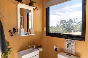 Une salle de bain avec un mur en bois, un lavabo blanc, un miroir et une fenêtre donnant sur des arbres.