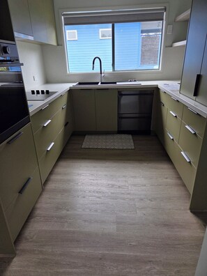 A compact kitchen with a built-in dishwasher, a window with blinds, and a patterned rug on the floor. - Holiday Rental Red Rocks Area Phillip Island (Cowes)