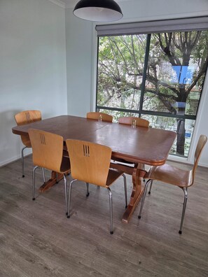 A dining area with a wooden table and chairs, a large window with a view of trees, and a hanging light fixture. - Holiday Rental Red Rocks Area Phillip Island (Cowes)