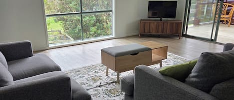 Ein geräumiges Wohnzimmer mit einer großen Schiebeglastür, die auf einen Balkon führt, einem Flachbildfernseher auf einem Holzschrank und zwei grauen Sofas.