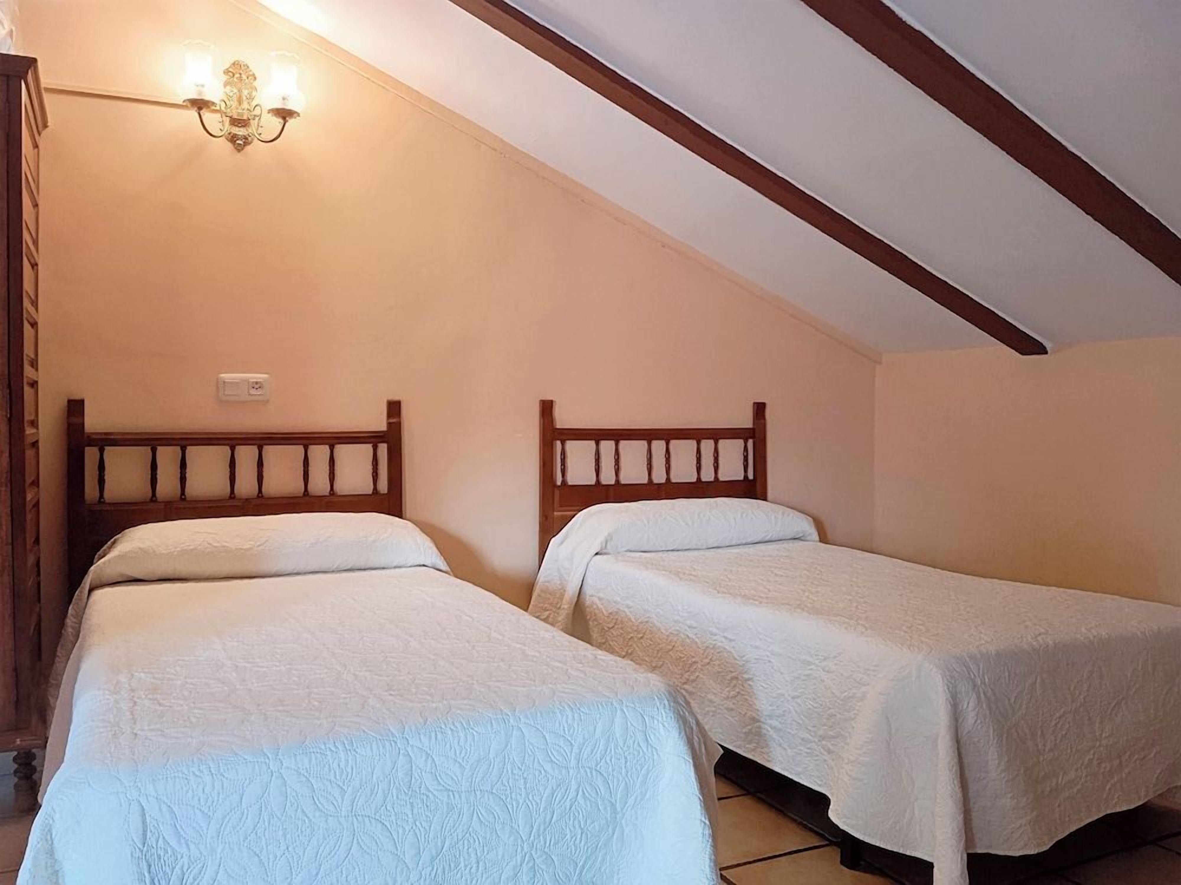 Una habitación con dos camas individuales, cabeceros de madera, una lámpara de pared y un techo inclinado con vigas de madera.