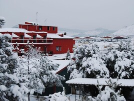 Un edificio rojo con múltiples ventanas y balcones, rodeado de árboles y colinas cubiertas de nieve al fondo.