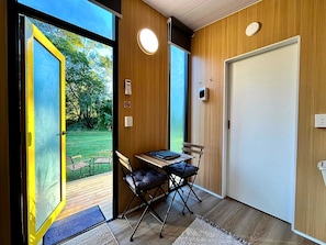 Ein Zimmer mit Holzboden, einem Tisch mit zwei Stühlen, einer Tür und einem Fenster mit Blick auf Grünflächen.