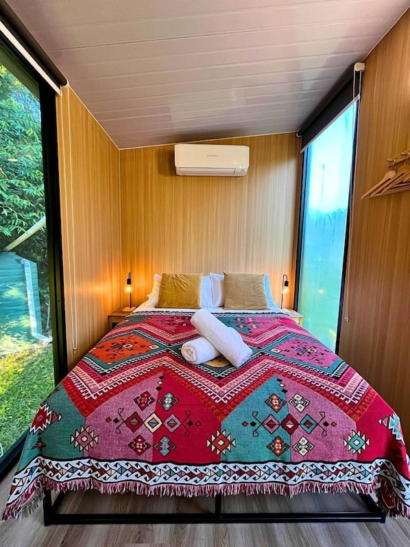 A modern bedroom with a large bed, a wooden ceiling, and a view of greenery outside.