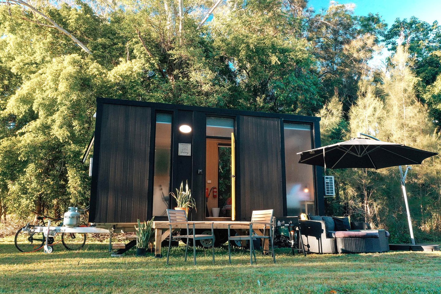 Une cabane moderne aux couleurs sombres, dotée d’une porte-fenêtre coulissante, d’une table avec des chaises, d’un parasol et d’un petit espace jardin.