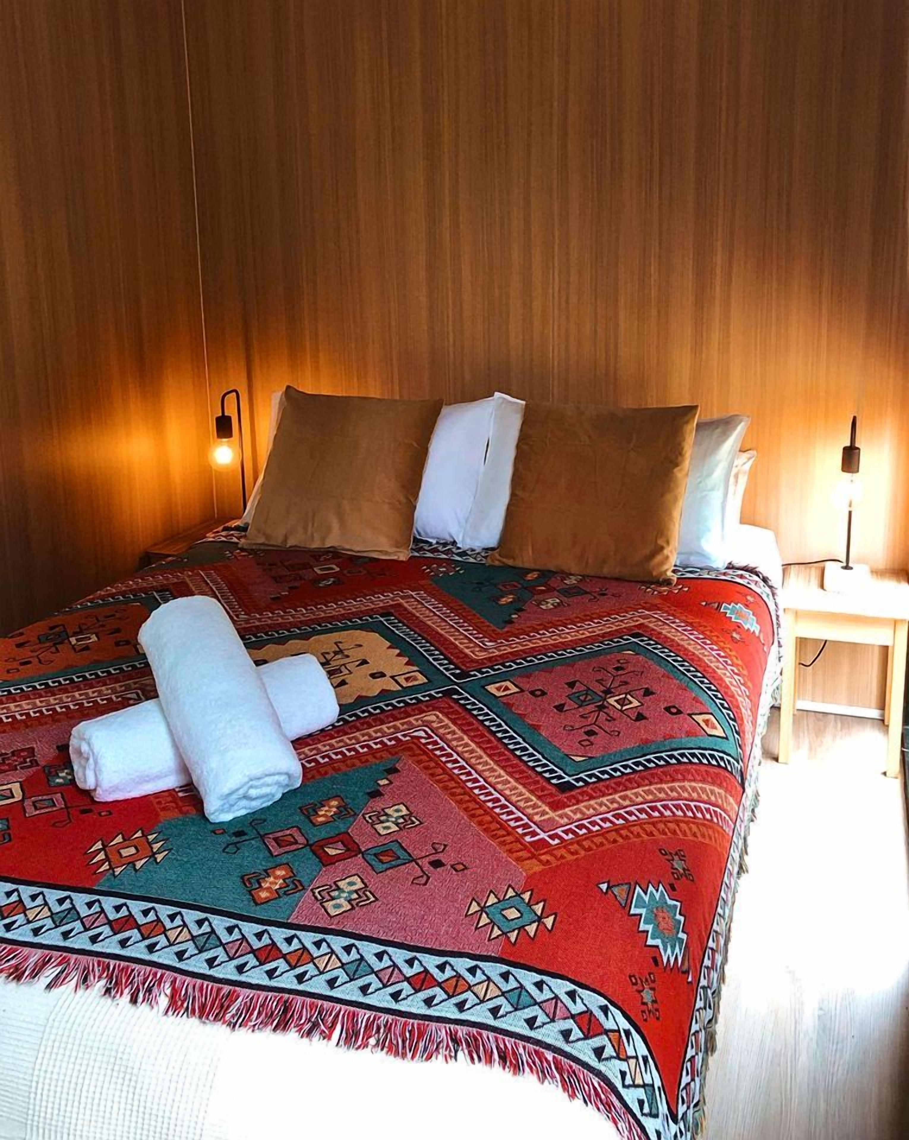 A hotel room with a wooden headboard, a patterned bedspread, two pillows, a bedside table with a lamp, and rolled towels on the bed.