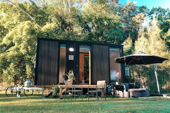 A modern, dark-colored cabin with a sliding glass door, a table with chairs, an umbrella, and a small garden area. - Tranquil Tiny Cabin Retreat with Stargazing near Sunshine Coast, Yandina QLD (Nambour)