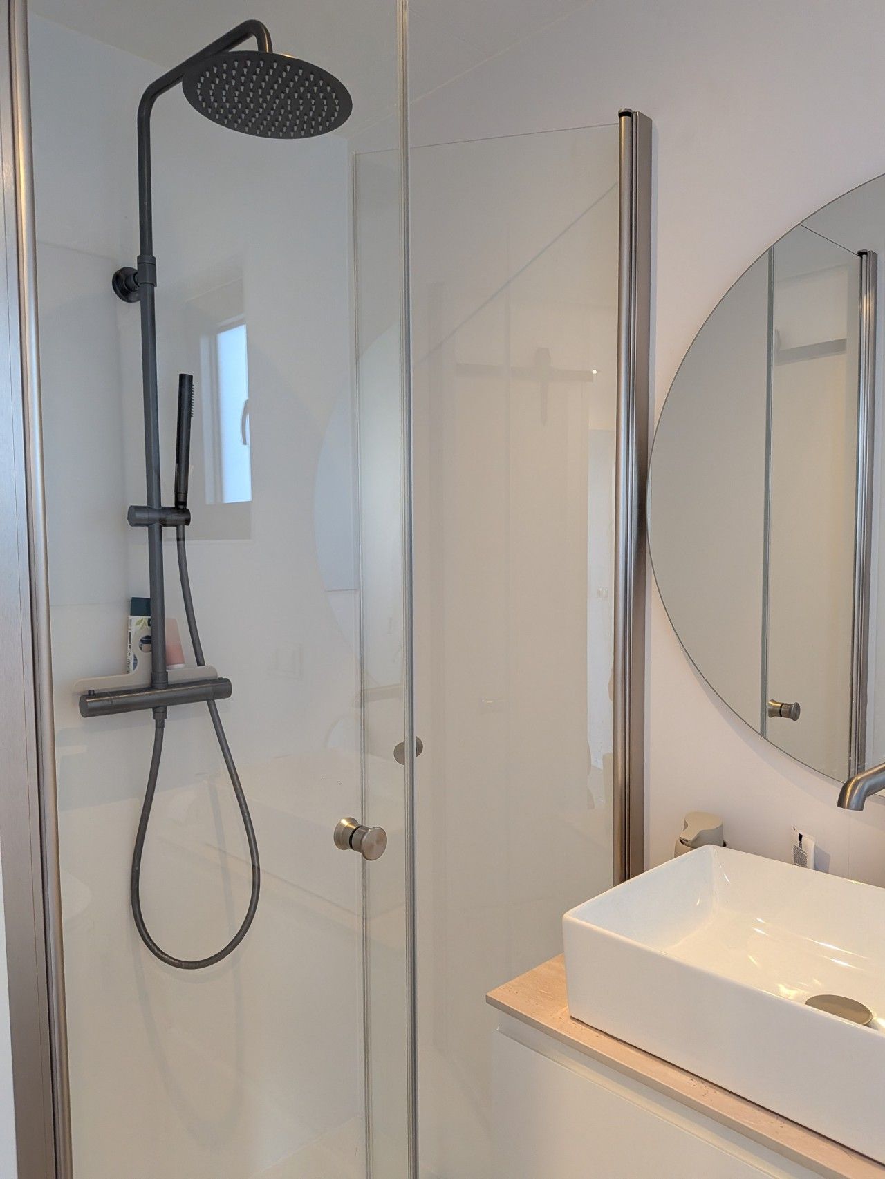 Een moderne badkamer met een douchecabine van glas, een witte wastafel en een ronde spiegel.