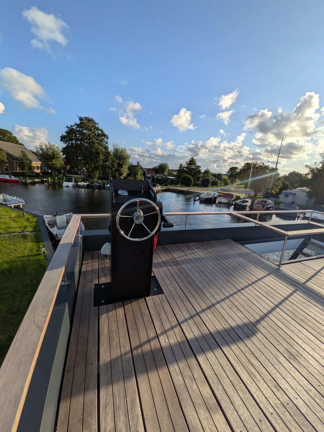 Ein Holzdeck mit Steuer, ruhigem Gewässer und Booten im Hintergrund.