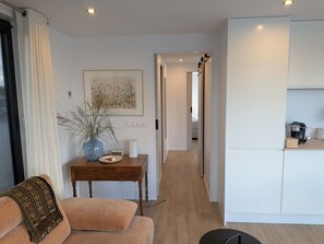 Ett modernt vardagsrum med en soffa, ett träbord och en vas med växter. En hall leder till ett kök med en kaffemaskin och skåp.