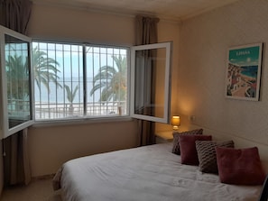 A hotel room with a bed, a window with a view of palm trees and the sea, a lamp, and a wall with a framed picture. - Beachfront Dos Nanitos, right on the beach. Private parking. Pets welcome! (San Pedro del Pinatar)