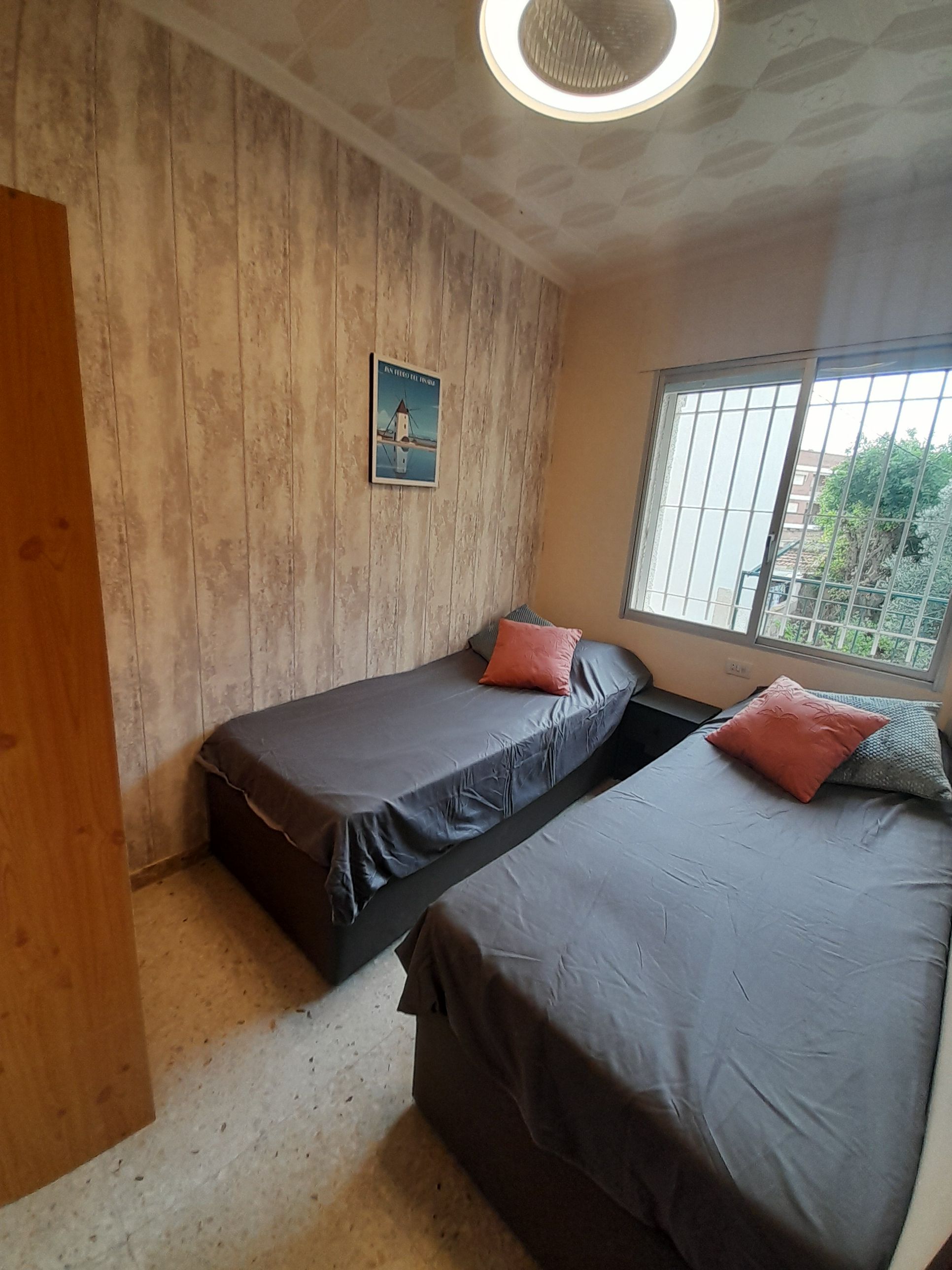 A room with two beds, a wooden wall, a poster, and a window with a view.