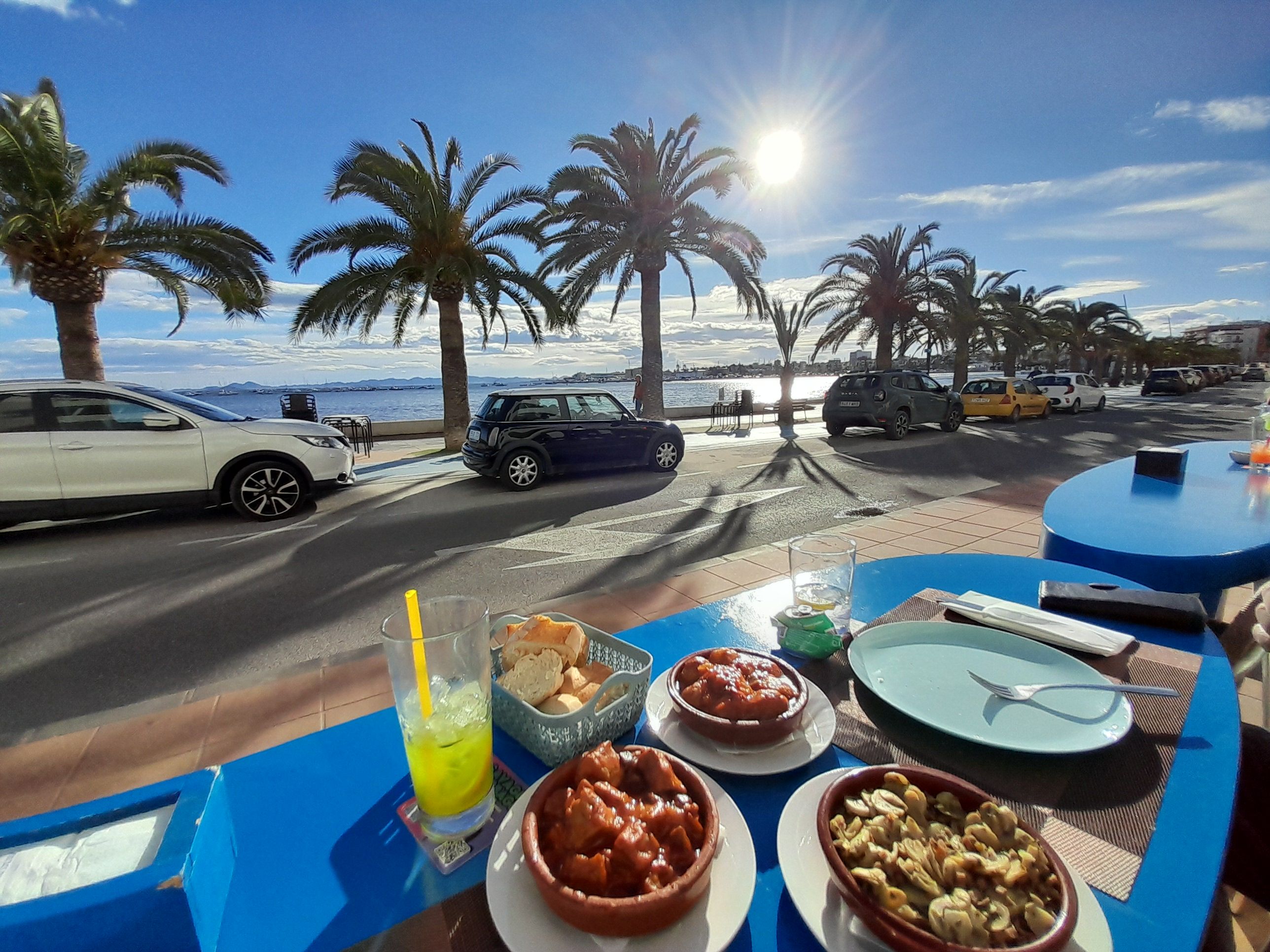 A table with food and drinks, overlooking a street with parked cars and palm trees.