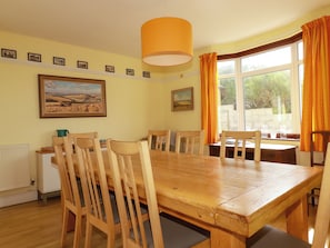 A dining area with a wooden table and chairs, a radiator, framed pictures on the wall, and a window with curtains. - Chy-an-lowena (Newquay)