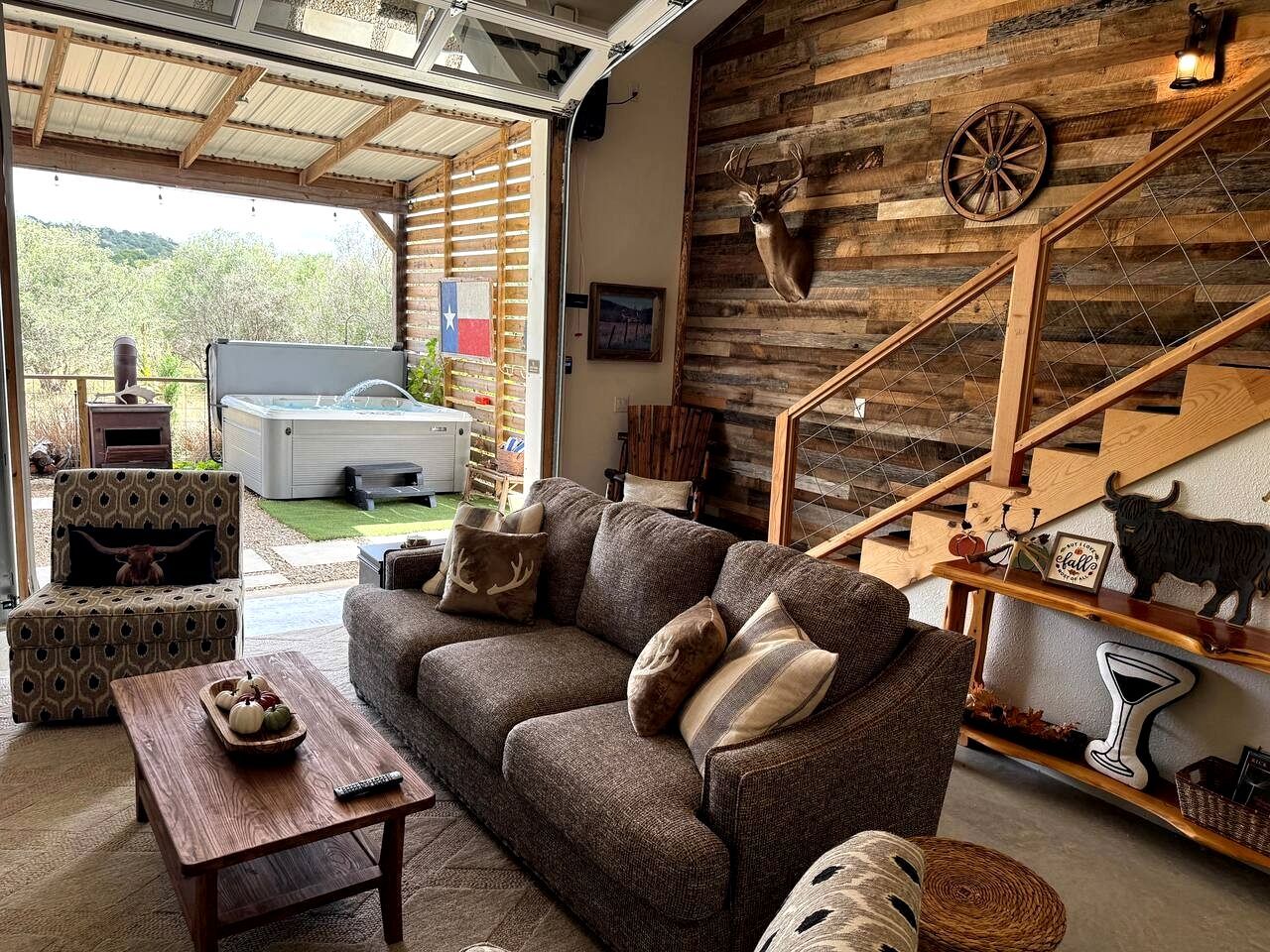 A cozy living room with a sofa, coffee table, and a fireplace. A wooden staircase and a wall-mounted deer head are visible.