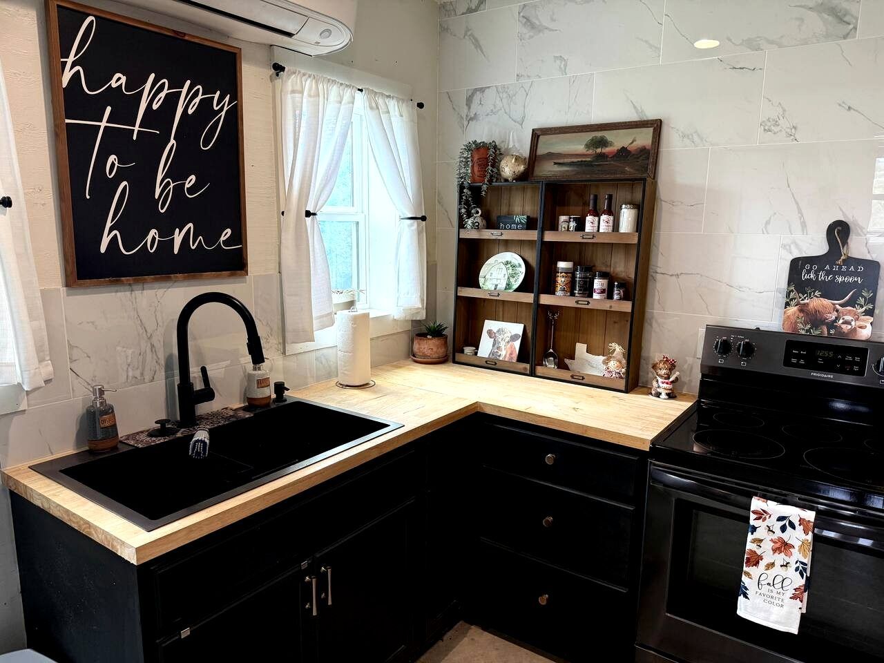 A kitchen with a black sink, wooden countertop, black cabinets, a stove, and a shelf with kitchen items.