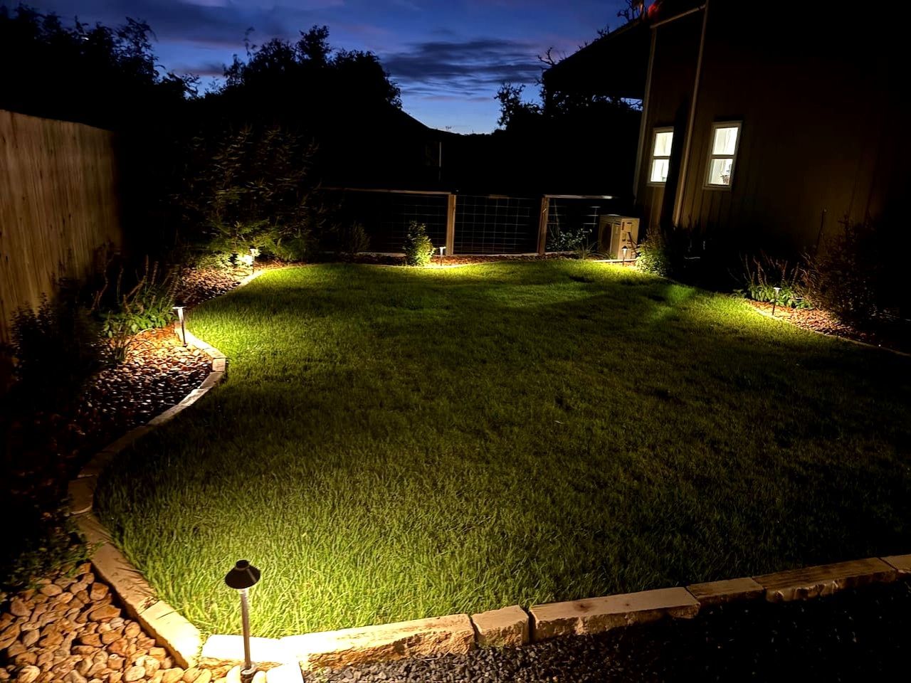 A well-lit backyard with a lawn, a wooden fence, and a house in the background.