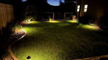 A well-lit backyard with a lawn, a wooden fence, and a house in the background.