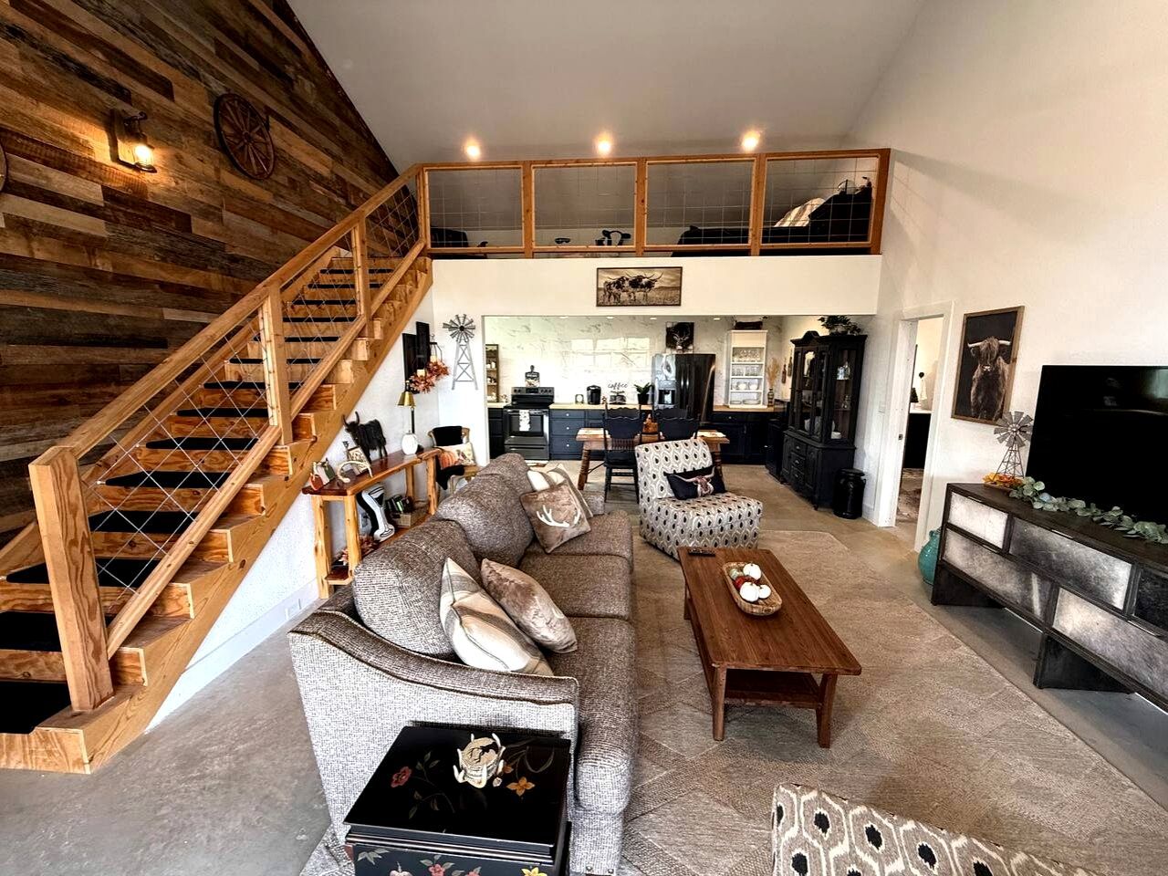 A spacious living area with a wooden staircase, a sofa set, a dining table, and a kitchen in the background.