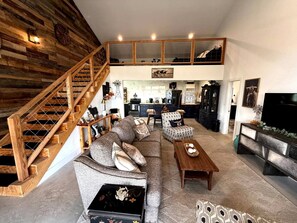 Een ruime woonkamer met een houten trap, een zithoek, een eettafel en een keuken op de achtergrond.