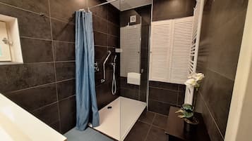 Een moderne badkamer met donkere tegels, een glazen douchecabine, een witte zeepdispenser en een potplant.
