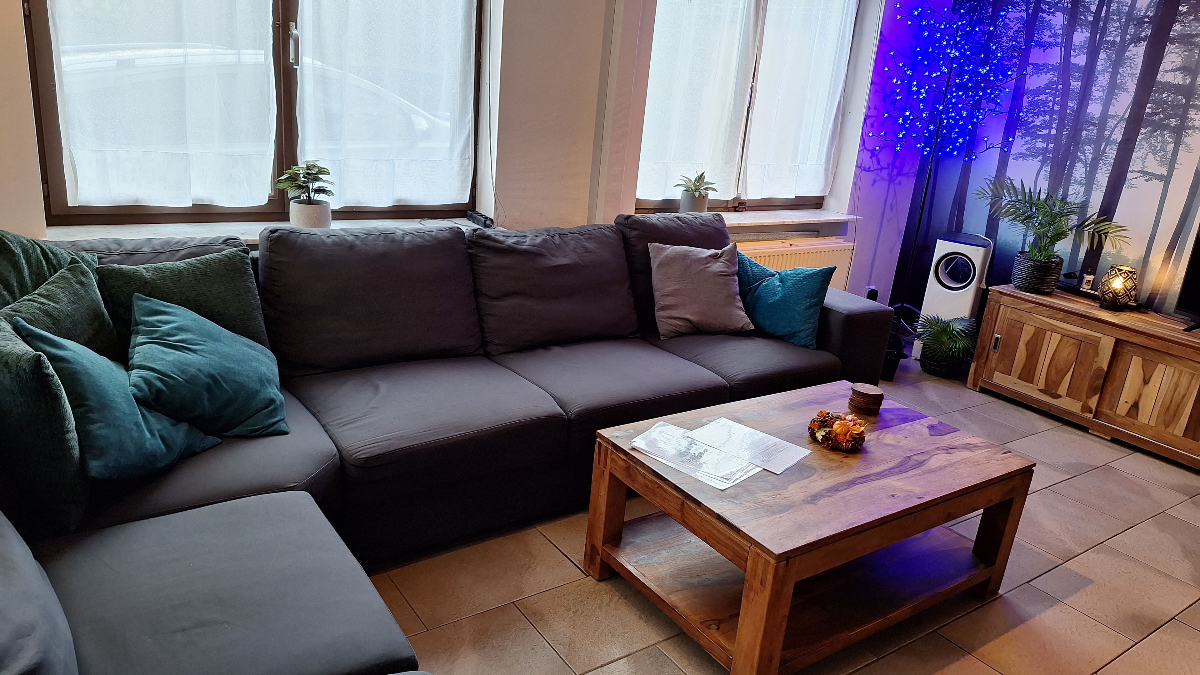 A modern living room with a dark brown sofa, teal cushions, a wooden coffee table, and a large window with sheer curtains.