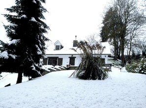 A snow-covered landscape with a house, trees, and a snow-covered bush.
