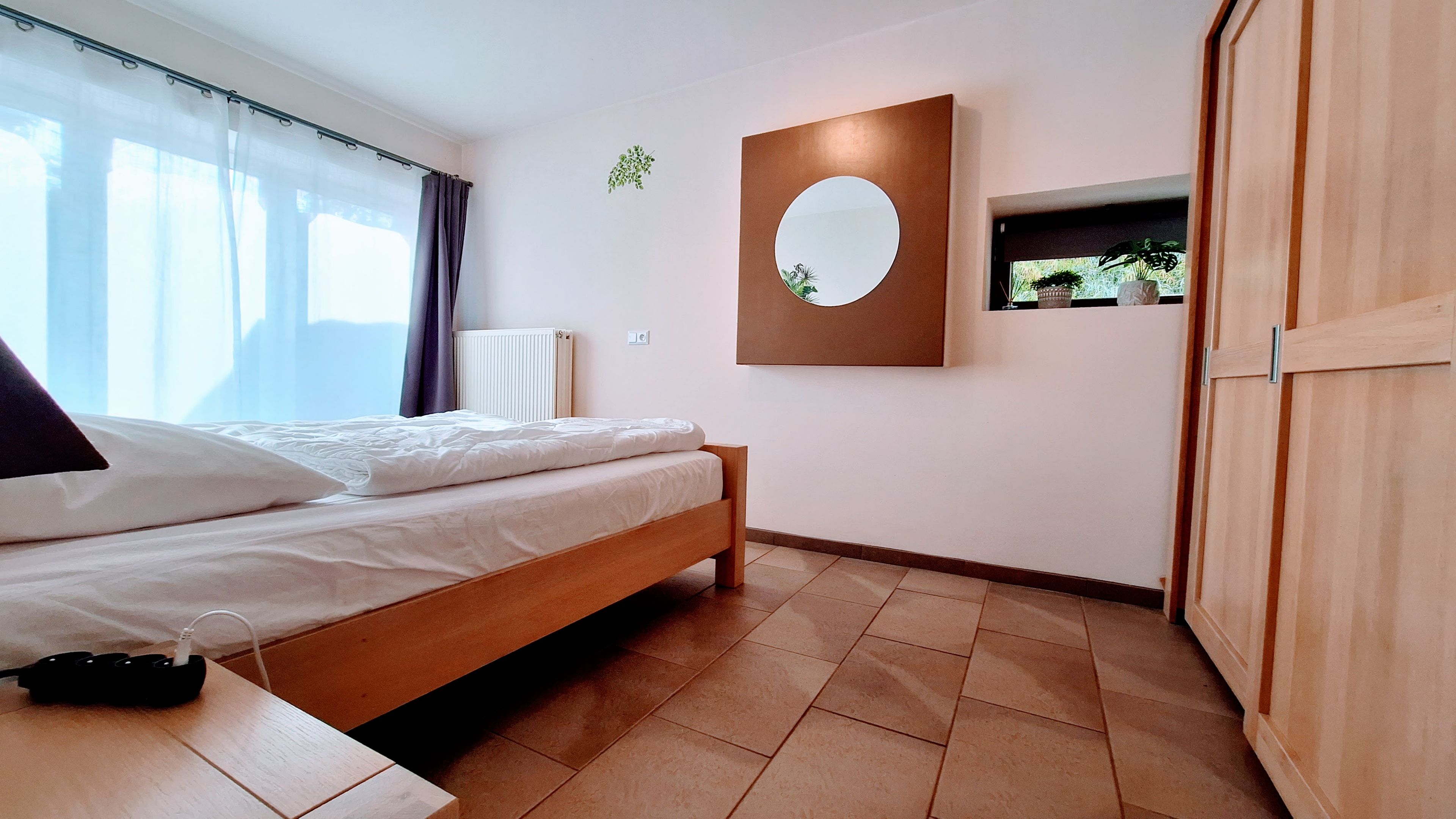 Een slaapkamer met een bed, nachtkastje met oplader, radiator, spiegel en een kledingkast.