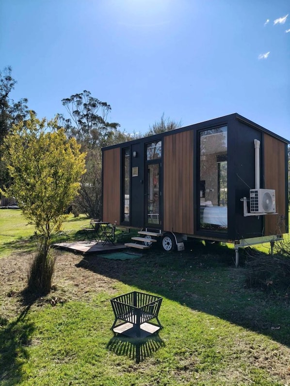 A modern, compact mobile home with a sliding door, a small patio area, and a visible air conditioning unit.