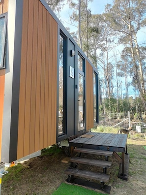 Un bâtiment moderne à l’extérieur en bois, doté d’une porte et d’un petit espace extérieur aménagé avec des sièges, accessible par quelques marches.