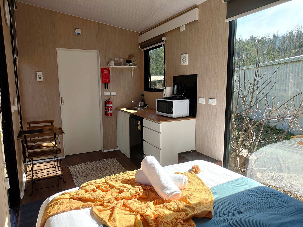A hotel room with a bed, a desk with a microwave, a small kitchenette, a fire extinguisher, and a view of the outdoors.