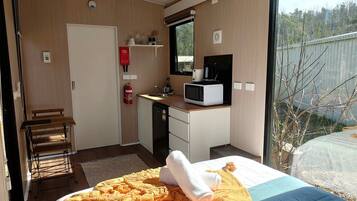 A hotel room with a bed, a desk with a microwave, a small kitchenette, a fire extinguisher, and a view of the outdoors.