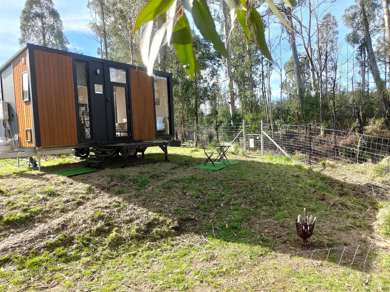Een kleine, moderne stacaravan met een houten buitenkant en een zwarte deur, gelegen in een grasrijk gebied met een omheining en bomen op de achtergrond.
